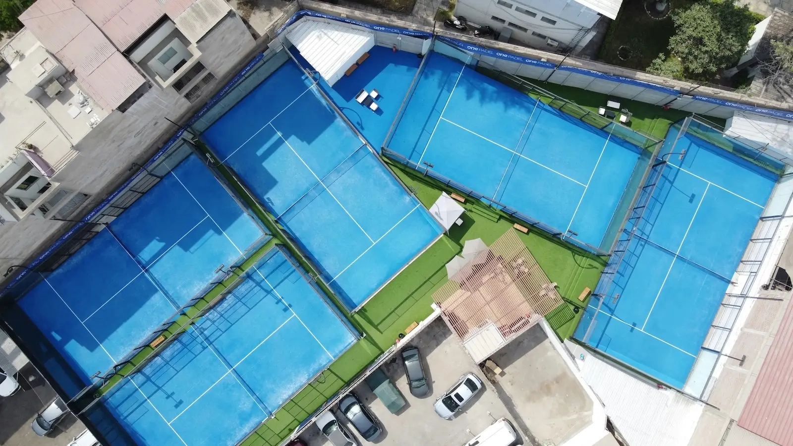 Een luchtfoto van vijf blauwe padelbanen, gelegen in een stedelijke omgeving met omliggende gebouwen en parkeerruimte.